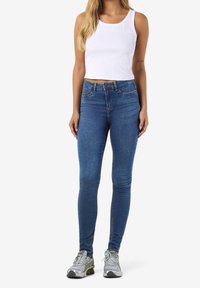 Femme portant un haut court blanc sans manches, un jean skinny taille haute bleu et des chaussures de course argentées, debout devant un fond blanc.
