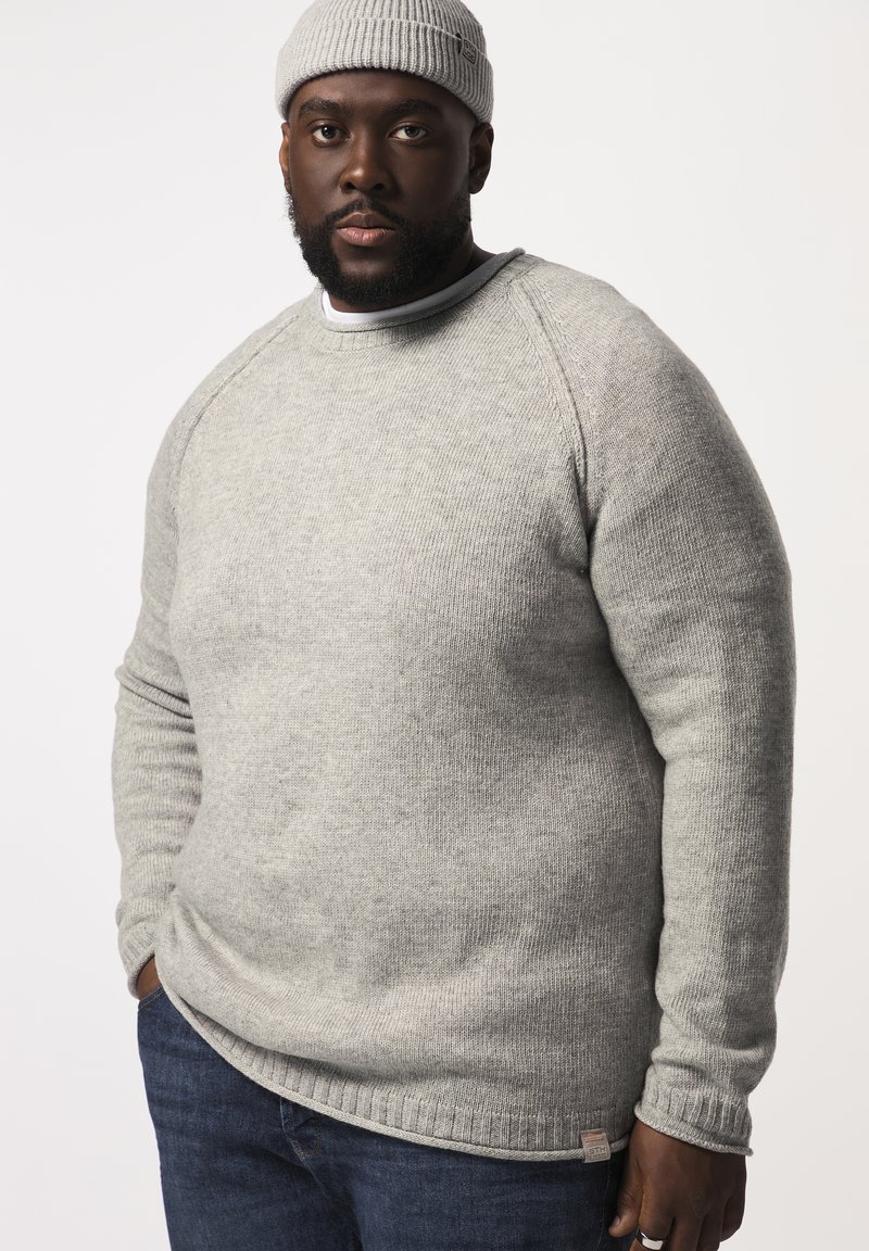 Maglione grigio lavorato a maglia con scollatura rotonda, polsini a coste e orlo dritto, abbinato a jeans blu scuro. Presenta un piccolo cartellino con il logo nella parte inferiore.