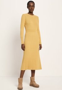 Eine Frau trägt ein senfgelbes, geripptes Midikleid mit langen Ärmeln und tanfarbenen Wildleder-Ankle-Boots mit Plateau, stehend vor einem einfarbigen Hintergrund.