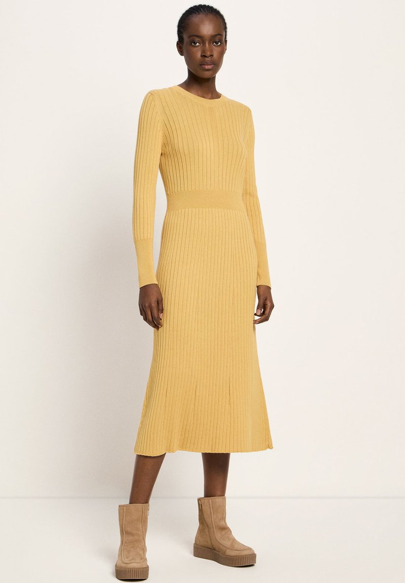 Eine Frau trägt ein senfgelbes, geripptes Midikleid mit langen Ärmeln und tanfarbenen Wildleder-Ankle-Boots mit Plateau, stehend vor einem einfarbigen Hintergrund.
