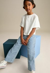 Vit kortärmad t-shirt, blå och vita randiga vida byxor, vita sneakers med röda accenter, poserar bredvid blå geometriska block.
