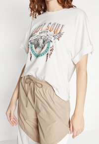 Bílé oversized tričko s grafickým designem, na němž je zobrazen orel, nošené s béžovými šortkami na stažení. Oba materiály vypadají měkce a ležérně.