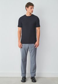 Sort mønstret t-shirt med korte ærmer, parret med lysegrå bukser og sorte sko, stående foran en ensfarvet hvid baggrund.
