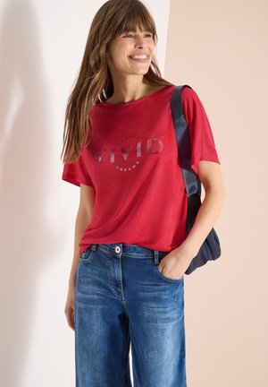Femme souriante portant un t-shirt rouge "Vivid Dreams", un jean bleu et un sac à bandoulière bleu marine, sur un fond neutre.