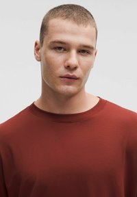 Rostfärgad, kortärmad skjorta med rund halsringning. Slät textur med en figurnära design. Enkel och ren estetik utan synliga mönster.