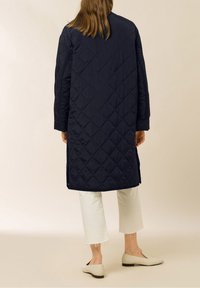 Abrigo largo acolchado azul marino con un ajuste relajado, que presenta un cuello alto y aberturas laterales, combinado con pantalones blancos recortados y zapatos beige sin cordones.