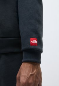 Temno siva majica s kapuco z ribanim manšetami, z rdečim kvadratnim logotipom z belim napisom, ki je nameščen na rokavu. Teksturirana tkanina.