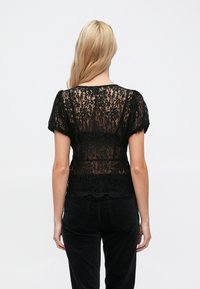 Čierny čipkovaný top s krátkymi nafúknutými rukávmi. Priehľadná textúra a kvetinové vzory. Lem s vlnkovaným okrajom, nosený s čiernymi nohavicami.