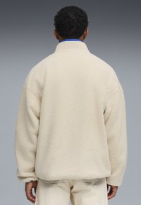 Beige fleece-pullove takki korkealla kaulalla, pudotetuilla hartioilla ja teksturoidulla pinnalla, yhdistettynä vaaleisiin housuihin.