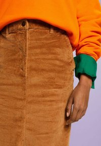 Brauner Cordrock mit vertikalen Rippungen, ausgestattet mit einem knöpfschnürenden Vorderteil. Darüber trägt sie einen orangefarbenen Pullover mit grünen Bündchen.