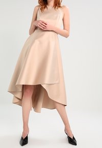 Beige ärmlös klänning med hög-låg fåll, gjord av ett slätt tyg. Har en figurnära bodice och utsvängd kjol. Svarta klackskor med spetsiga tår.