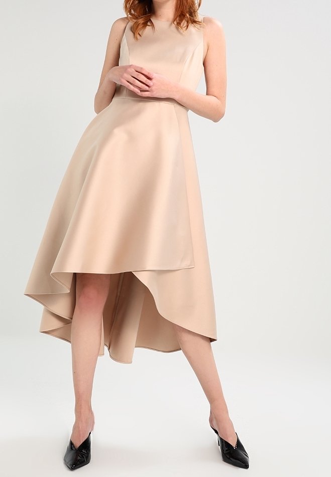 Beige ärmlös klänning med hög-låg fåll, gjord av ett slätt tyg. Har en figurnära bodice och utsvängd kjol. Svarta klackskor med spetsiga tår.