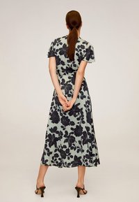 Robe midi florale de couleur vert clair avec un motif floral foncé, à manches courtes et ourlet évasé. Le modèle se tient debout avec les mains jointes dans le dos.