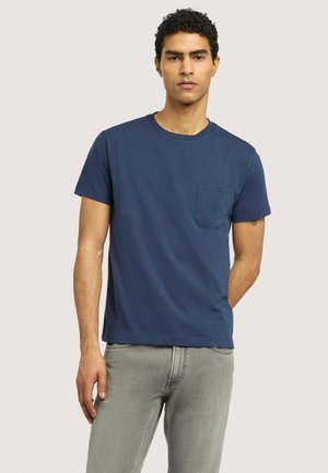 Mężczyzna o kręconych włosach, ubrany w granatowy t-shirt z kieszonką i jasnoszare dżinsy, stoi na tle gładkiego jasnego tła.