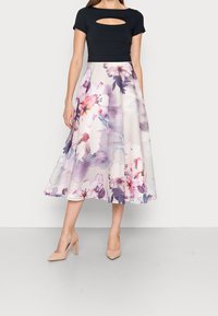 Femme portant un top noir à manches courtes avec un décolleté en forme de trou de serrure et une jupe midi plissée blanche ornée de grands motifs floraux roses et violets.