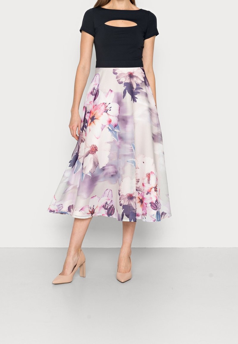 Femme portant un top noir à manches courtes avec un décolleté en forme de trou de serrure et une jupe midi plissée blanche ornée de grands motifs floraux roses et violets.