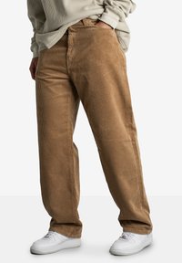 Osoba w luźnych brązowych spodniach sztruksowych, beżowym swetrze i białych trampkach, stojąca z jedną ręką w kieszeni.