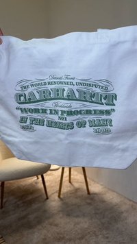Bela torbica iz platna z zelenim besedilnim grafičnim dizajnom. Besedilo vključuje »Carhartt« in »Work in Progress« z dekorativnimi poudarki.