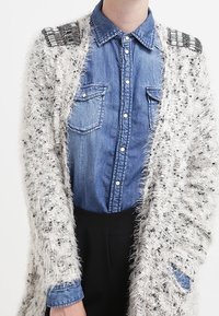 Vit fluffig cardigan med en strukturerad stickning, silverdetaljer på axlarna, bärs över en blå denimskjorta med två bröstfickor.