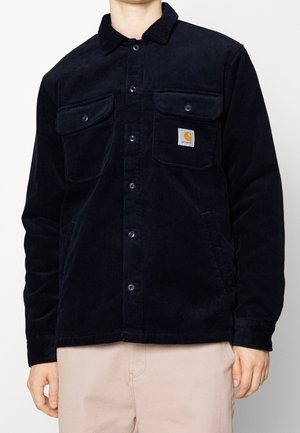 Mand iført sort manchetjakke med knapper og to brystlommer, Carhartt-logo på venstre lomme og lys beige bukser.
