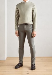 Felpa verde chiaro, camicia bianca con colletto sotto, pantaloni grigi slim-fit e scarpe di pelle marroni; texture morbide, design minimalista.