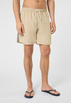 Uomo che indossa pantaloncini beige con coulisse e striscia laterale, e infradito blu navy, in piedi contro uno sfondo chiaro e uniforme.