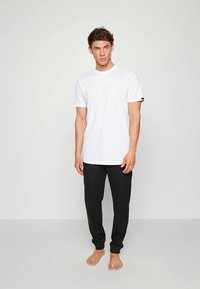 Hvid bomulds t-shirt med rund hals og korte ærmer, parret med sorte sweatpants med afslappede ankler. Modellen står barfodet.