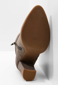 Hnědá semišová kotníková bota s texturovanou podrážkou, mírně zúženým podpatkem, bočním zipem a minimalistickým designem. Pohled ze šikmého úhlu proti zdi.