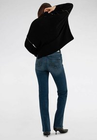 Mujer con vaqueros azul oscuro, tacones negros de punta afilada y un suéter negro holgado, de pie de espaldas con la mano levantada hacia la cabeza.