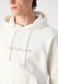 Sudadera blanca con cordón, que presenta un logo bordado de "CK" y el texto "Calvin Klein Jeans". Tejido suave y diseño minimalista.