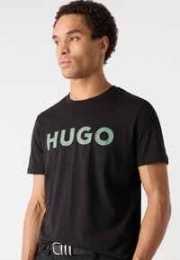 Fekete pamut póló, nagy zöld "HUGO" logóval a mellkasán, kerek nyakkal és rövid ujjakkal. Sima textúra és laza fazon.