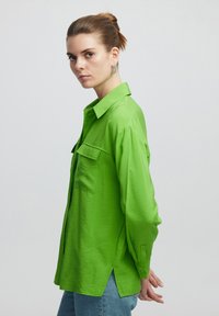 Limonkowa, długa koszula z luźnym krój, z wyraźnym kołnierzykiem i przednimi kieszeniami. Teksturowana tkanina, boczne rozcięcia i mankiety na guziki.