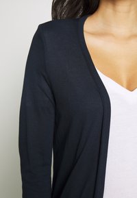 Cardigan marine à texture côtelée, à l'avant ouvert, avec des manches longues, superposé sur un t-shirt blanc à col en V.