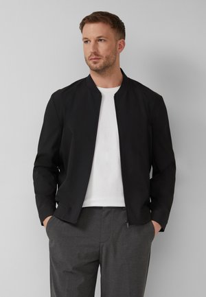 Homme portant une veste noire zippée par-dessus une chemise blanche et un pantalon gris, les mains dans les poches, regardant vers la gauche sur un fond gris uni.