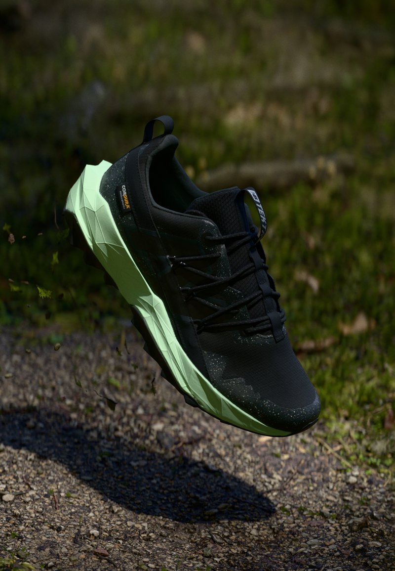 Schwarz-grüner Trailrunning-Schuh mit strukturierten Sohlen in der Luft über einem Schotterweg und verschwommenem, grünem Hintergrund.