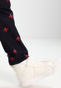 Jambe portant un pantalon noir à motifs fleur-de-lys rouges et des baskets montantes blanches à lacets sur un fond blanc uni.
