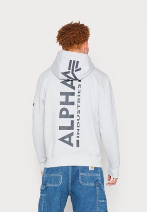 Mężczyzna z kręconymi rudymi włosami, ubrany w biały kaptur z dużym pionowym napisem „ALPHA INDUSTRIES” na plecach oraz niebieskie dżinsy.