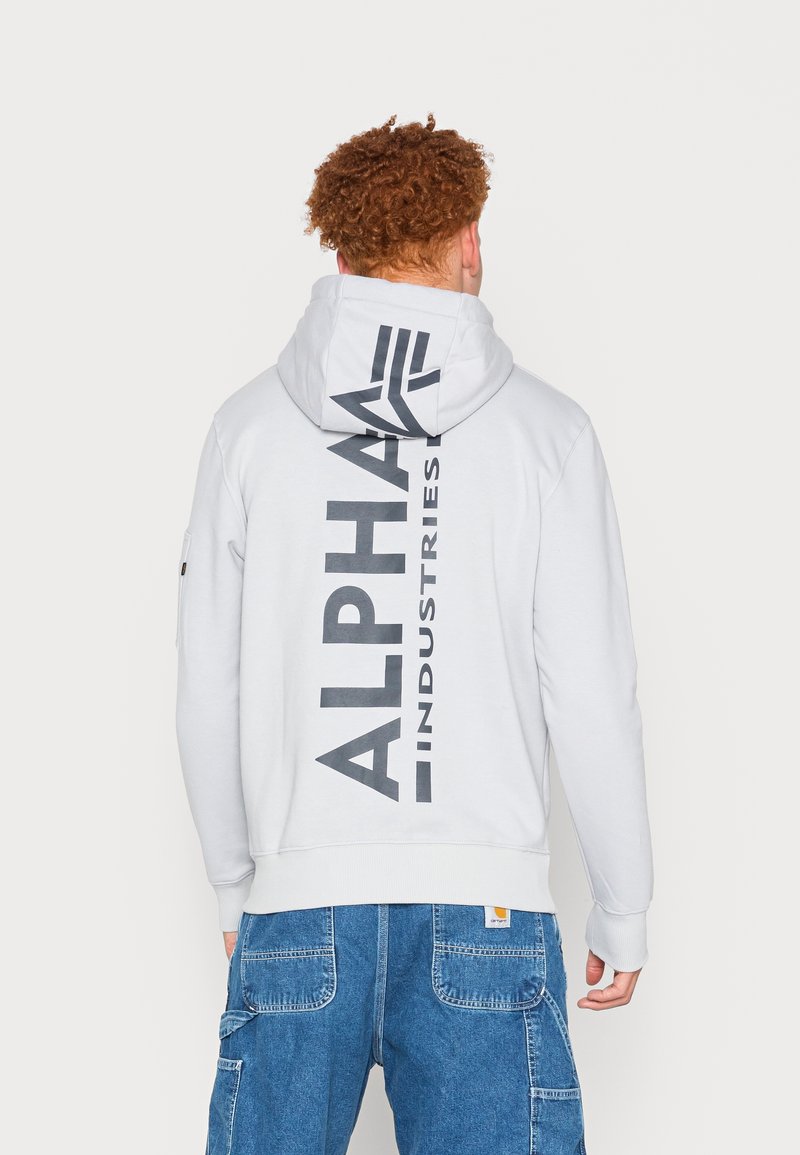 Homme aux cheveux roux bouclés portant un sweat à capuche blanc avec un grand texte vertical « ALPHA INDUSTRIES » dans le dos et un pantalon en jean bleu.