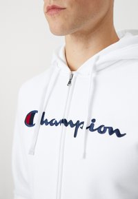 Hvit hettejakke med glidelås laget av mykt stoff. Har marineblå brodert "Champion"-logo og snor. Glatt tekstur med en uformell design.