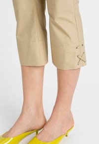 Pantaloni beige a coste con risvolto, caratterizzati da dettagli in pizzo e occhielli in metallo, abbinati a mules gialli lucidi che evidenziano il design elegante delle calzature.