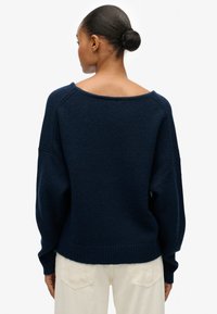 Granatowy, oversize sweter z szerokim dekoltem, teksturowanym materiałem, prążkowanym wykończeniem i opuszczonymi szwami ramion, zestawiony z kremowymi spodniami.