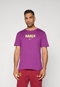 Lilla bomulds t-shirt med "BARÇA" i gul, med et lille Nike-logo under. Modellen har maroon bukser på med logodetalje.