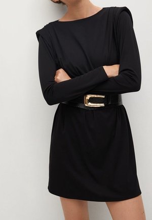 Femme portant une robe noire à manches longues avec des épaules rembourrées et une large ceinture noire avec une grande boucle dorée, bras croisés.