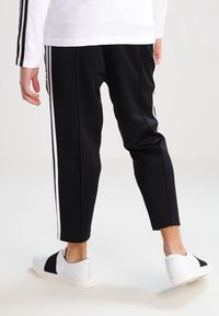 Osoba nosící černé teplákové kalhoty s bílými pruhy po stranách a bílé slip-on tenisky s černými pásky, stojící na jednoduché bílé podlaze.