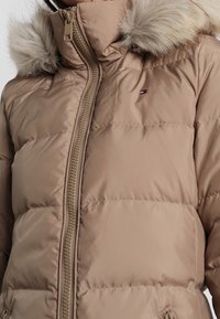 Béžová péřová bunda s vysokým límcem, zdobená umělou kožešinou na kapuci. Obsahuje přední zip a malé logo na levé hrudi.