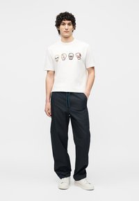 Νέος άνδρας με σγουρά μαλλιά που φοράει λευκό μπλουζάκι με τέσσερα πολύχρωμα αποτυπώματα κρανίων, μαύρο φαρδύ παντελόνι και λευκά αθλητικά παπούτσια.