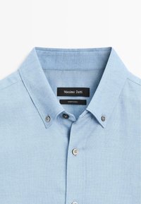 Hellblaues Button-Up-Hemd aus weichem Stoff mit einem Kentkragen, zwei Knöpfen am Kragen und einem schwarzen Markenetikett innen.