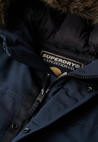 Marineblaue isolierte Jacke mit einer Kapuze aus Kunstfell, ausgestattet mit einem Reißverschluss, Druckknöpfen und einem Stoffetikett von Superdry Expedition Co.