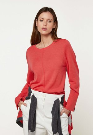 Frau mit langen braunen Haaren, die einen roten Pullover und weiße Hose trägt, mit den Händen in den Taschen stehend vor einem einfarbigen hellen Hintergrund.