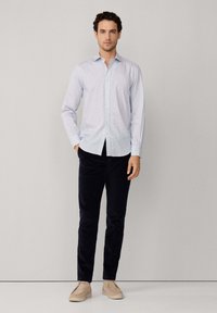 Hombre de pie contra un fondo blanco, vestido con una camisa de cuadros celeste, pantalones negros y zapatos casuales beige.